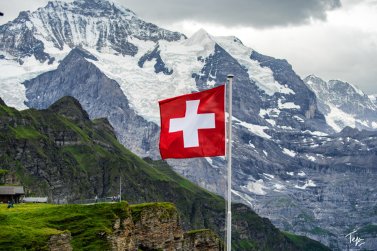 a flag with a white cross on a pole in front of a snowy mountain