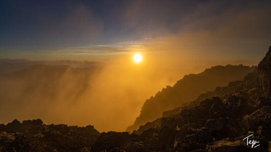 a sun shining through clouds over a rocky mountain