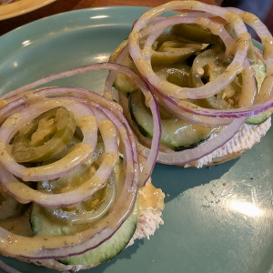 The image shows two open-faced sandwiches on a green plate. Each sandwich is topped with slices of cucumber, pickles, and red onion rings, drizzled with a creamy dressing. The sandwiches appear to be on a base of bread or a similar item. Another plate with a similar sandwich is partially visible in the background.