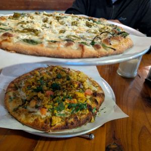 The image shows two pizzas on metal stands. The top pizza has a thin crust with toppings including spinach, jalapeños, and crumbled cheese. The bottom pizza has a thicker crust with toppings such as diced tomatoes, cilantro, and what appears to be seasoned cauliflower. A person is partially visible in the background, sitting at a wooden table.