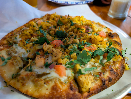 A close-up of a pizza topped with various ingredients, including diced tomatoes, herbs, and possibly pieces of chicken or paneer. The crust is golden brown and slightly crispy. The pizza is placed on a piece of parchment paper, and there are drinks in the background on a wooden table.