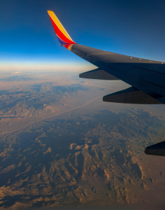 Aerial view from an airplane window showing a vast desert landscape with rugged mountains and valleys. The airplane wing is visible in the foreground, with a clear blue sky above. The terrain below is illuminated by sunlight, highlighting the textures and contours of the land.