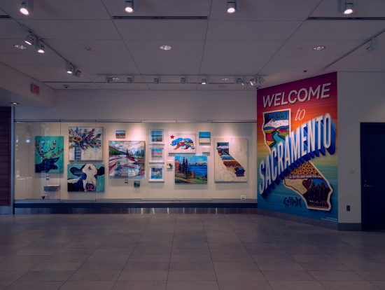 The image shows an art display in a gallery or public space. On the left, there are several colorful paintings featuring landscapes, animals, and abstract designs. On the right, there is a large mural with the text "Welcome to Sacramento" in bold letters, accompanied by a stylized map of California with various illustrations. The space is well-lit with overhead lights, and the floor is tiled.