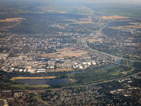Aerial view of a cityscape featuring a river winding through the area. The city is densely packed with buildings, roads, and industrial areas. There are patches of greenery and parks along the riverbanks. The landscape extends into the distance with a mix of urban and rural areas, including fields and open land.