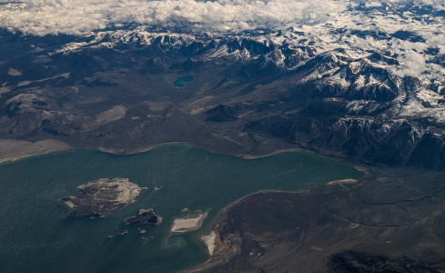 Aerial view of a large lake with several small islands in the foreground. The water is a deep greenish-blue color. Surrounding the lake is a rugged landscape with dark, barren terrain. In the background, there are snow-capped mountains under a partly cloudy sky. The scene is expansive and dramatic, showcasing a mix of water, land, and mountains.