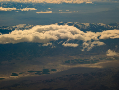 Aerial view of a mountainous landscape partially covered by clouds. Snow-capped peaks are visible in the background, while the foreground shows a series of circular fields or irrigation plots on a flat plain. The scene is bathed in soft, diffused light.