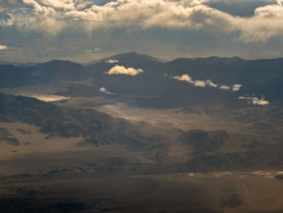 Aerial view of a vast desert landscape with rugged mountain ranges. The terrain is mostly barren with some scattered clouds casting shadows on the ground. The sky is partly cloudy, adding depth to the scene.