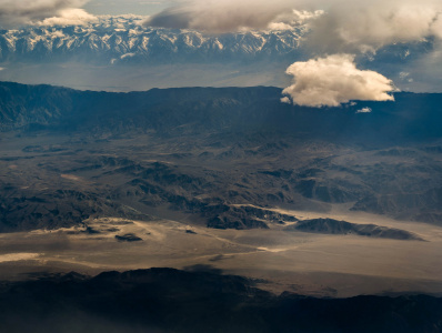 Aerial view of a vast desert landscape with rugged, dark brown mountains and sandy plains. In the background, there are snow-capped mountains partially covered by clouds. The sky is mostly cloudy, casting shadows over the terrain.