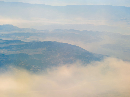 Aerial view of a mountain range partially obscured by clouds and mist. The landscape features rugged terrain with peaks and valleys, and the atmosphere appears hazy, giving a soft, ethereal quality to the scene. The sky above is light and blends with the misty horizon.