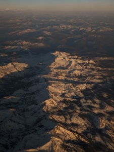 Aerial view of a mountain range with snow-covered peaks and ridges. The landscape is bathed in warm, golden light, likely from a sunrise or sunset, casting long shadows across the terrain. The sky above is clear with a hint of haze near the horizon.