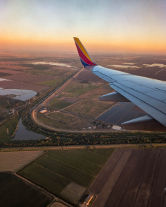 Aerial view from an airplane window showing a wing and winglet with a colorful design. Below, there are expansive agricultural fields, roads, and water bodies. The sky is clear with a warm, orange hue, suggesting a sunrise or sunset.