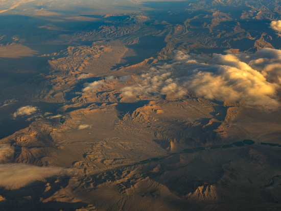 Aerial view of a vast desert landscape with rugged terrain and mountain ranges. The scene is partially covered by fluffy clouds casting shadows on the ground. The lighting suggests a sunrise or sunset, highlighting the textures and contours of the land.