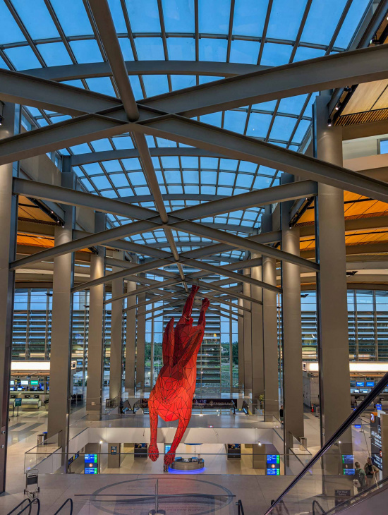 The image shows the interior of an airport terminal with a large, red geometric sculpture of a rabbit suspended from the ceiling. The terminal features a modern design with a high, glass-paneled roof and metal beams. There are check-in counters and digital screens visible on the lower level. The overall atmosphere is spacious and well-lit.