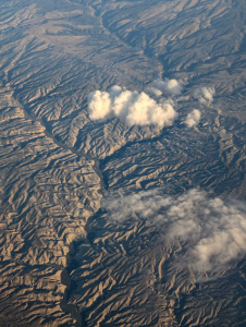 Aerial view of a rugged, mountainous landscape with deep valleys and ridges. Several fluffy white clouds are scattered above the terrain, casting shadows on the ground below. The lighting suggests a late afternoon or early morning setting, highlighting the contours of the landscape.