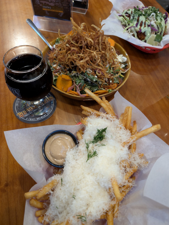 The image shows a table with various food items. In the foreground, there is a basket of fries topped with grated cheese and garnished with herbs, accompanied by a small cup of dipping sauce. To the left, there is a glass of dark beer on a coaster. In the background, there is a plate with a salad topped with crispy fried onions and a basket containing a dish with shredded cabbage and a lime wedge.