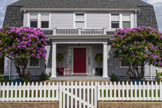 A two-story house with gray siding and a dark gray roof. The front features a red door with white trim, flanked by two large windows. Two purple-flowered trees are on either side of the entrance. A small porch with two red chairs and potted plants is visible. A white picket fence runs along the front of the property.