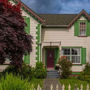 A charming house with a colorful exterior featuring green and pink trim. The front door is maroon, and there are green shutters on the windows. A white picket fence borders the front yard, which has lush greenery and a large tree with dark red leaves. The sky above is cloudy.