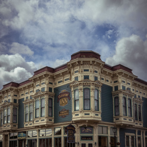 The image shows a large, ornate Victorian-style building with intricate architectural details. The building is painted in shades of blue and cream, with multiple large windows and decorative moldings. Signs on the building indicate it is the "Victorian Inn" offering lodging and a restaurant. The sky above is filled with dramatic clouds.