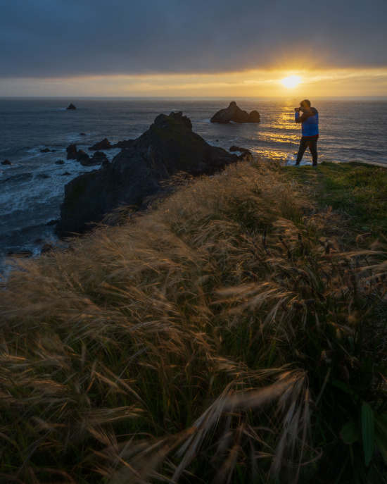 A person is standing on a grassy cliff, taking a photo of the ocean at sunset. The sun is setting on the horizon, casting a warm glow over the water and rocky outcrops. The sky is partly cloudy, and the foreground features tall grasses swaying in the breeze.
