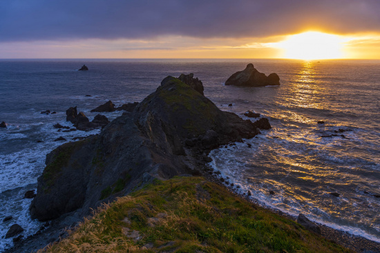 A scenic view of a rocky coastline at sunset. The sun is setting on the horizon, casting a warm golden light over the ocean. The foreground features grassy cliffs and rugged rocks, with waves crashing against them. The sky is partly cloudy, with a mix of dark and illuminated clouds.