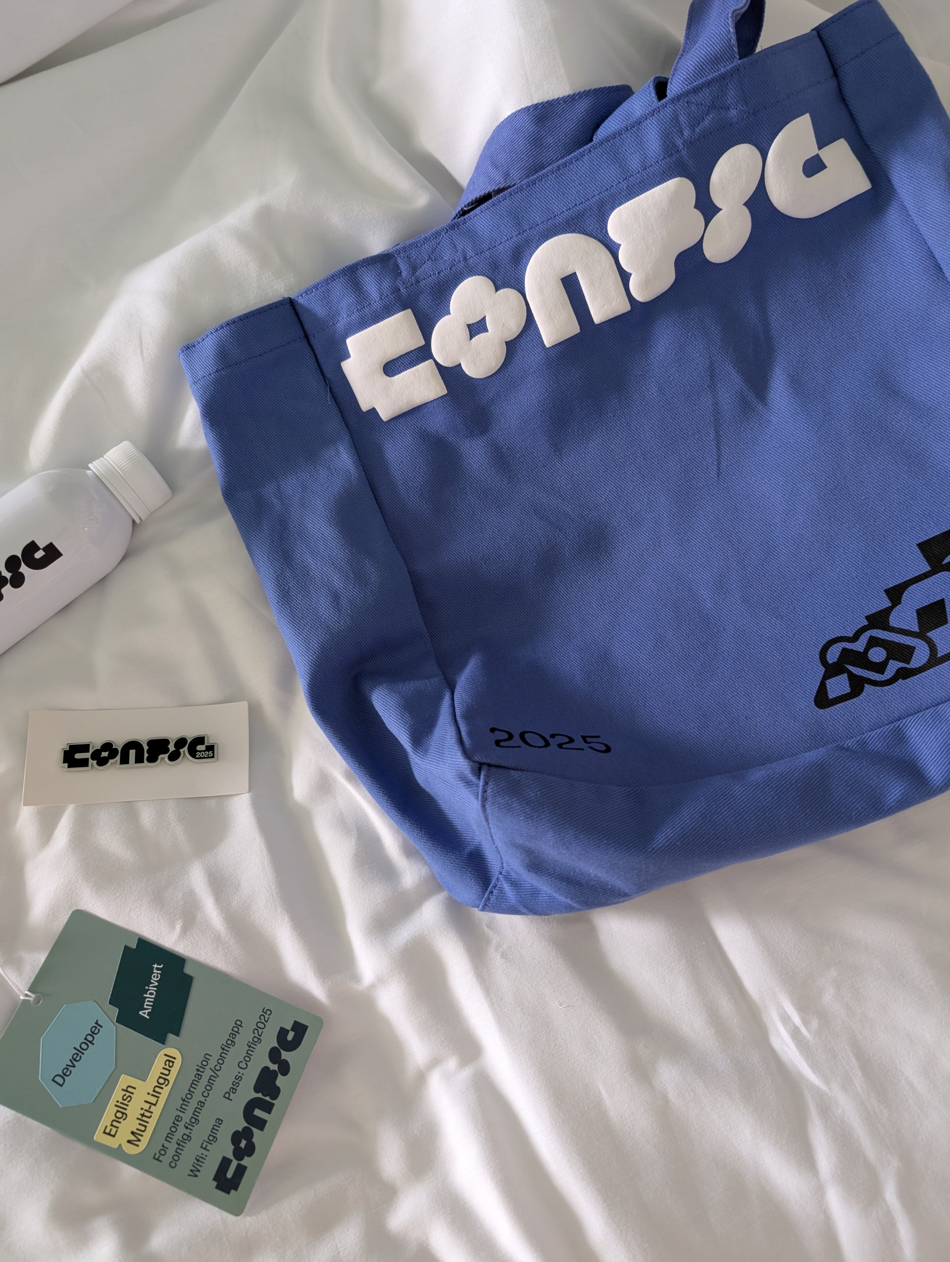 A blue tote bag with white and black text and designs is placed on a white surface. The bag has "2025" printed on it. Nearby, there is a white bottle with black text, a sticker with similar text, and a lanyard with a card that has various labels such as "Developer," "English," "Multilingual," and "Ambivert." The card also mentions WiFi and a hashtag.