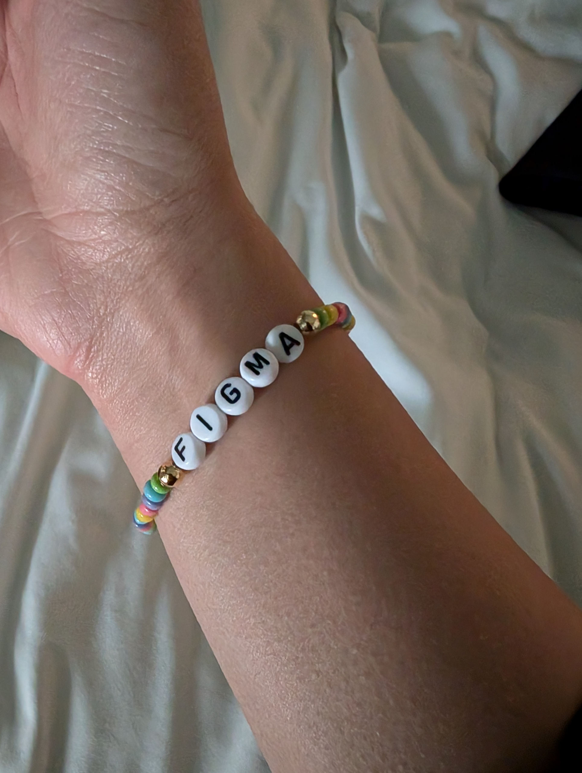 A close-up of a person's wrist wearing a beaded bracelet. The bracelet features white beads with black letters spelling "FIGMA" and is interspersed with colorful and gold beads. The background is a light-colored fabric.