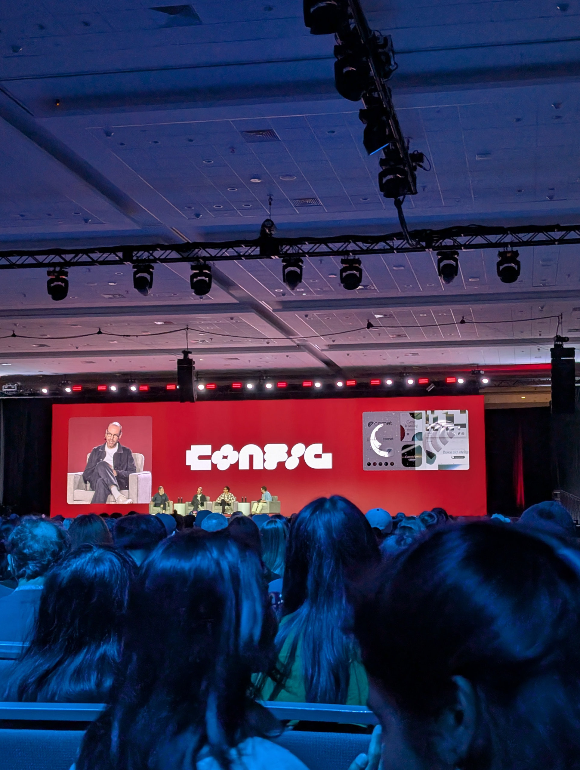 The image shows a large conference room filled with attendees. On stage, there is a panel of speakers seated in front of a large red screen displaying the word "Config" and various graphics. The room is dimly lit, with spotlights illuminating the stage. The audience is focused on the presentation.