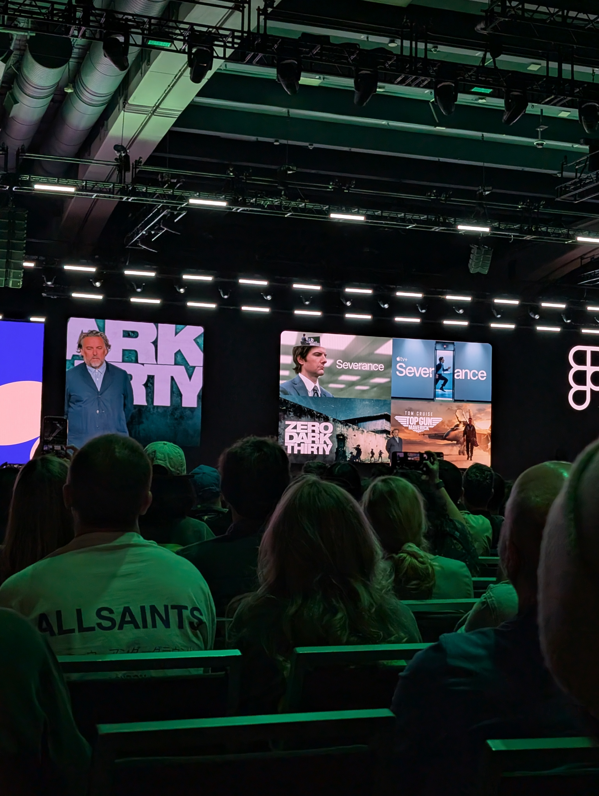 The image shows a large indoor event with a stage and an audience. On the stage, there are multiple screens displaying images and text related to various media titles, including "Severance," "Zero Dark Thirty," and "Top Gun: Maverick." A person is standing on the stage, and the audience is seated, watching the presentation. The venue has visible lighting and ceiling structures.