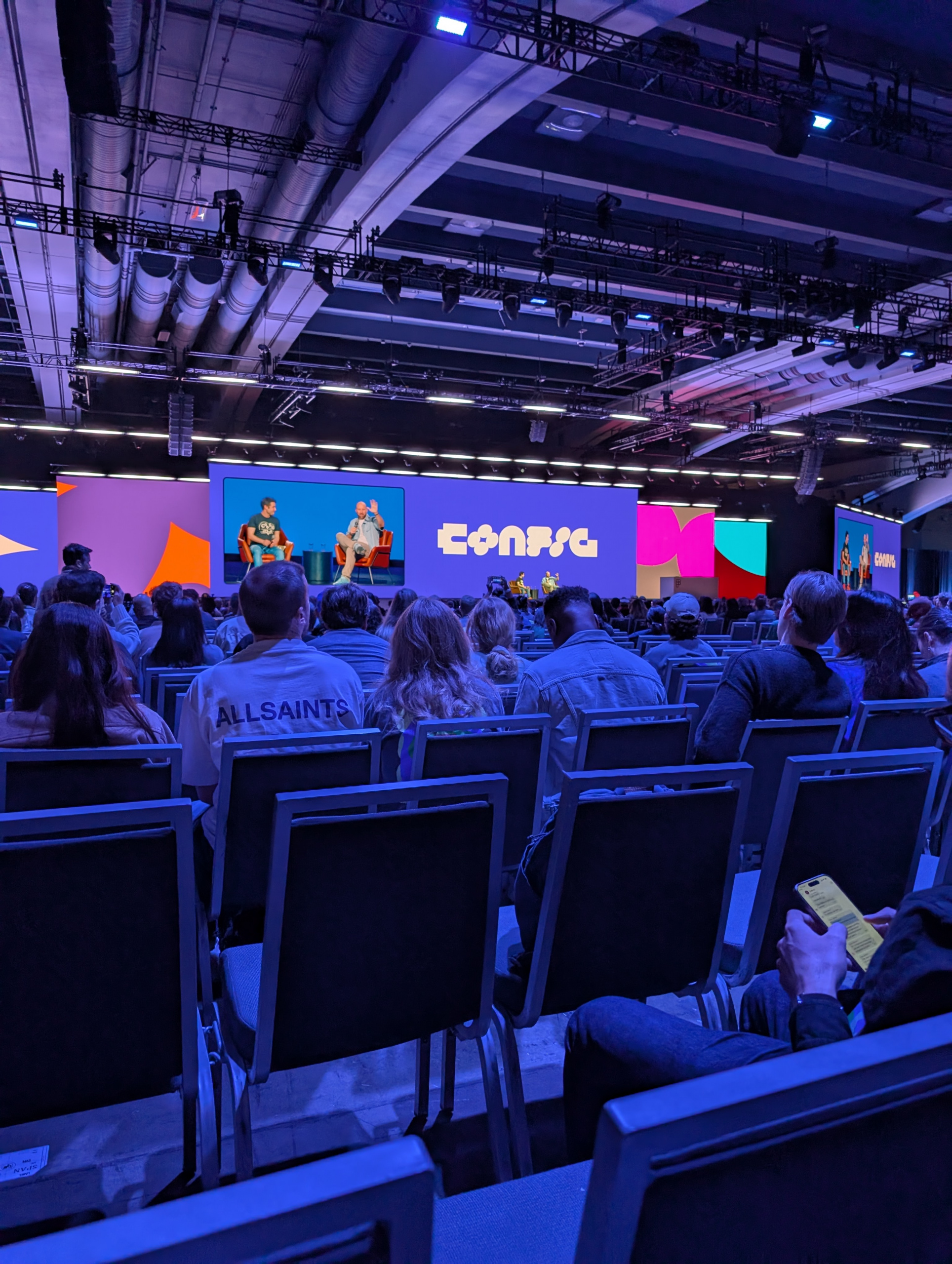 The image shows a large conference room filled with people seated in rows of chairs, facing a stage. On the stage, there are two people sitting and talking, with a large screen behind them displaying their image. The screen also has the word "Config" in bold letters. The room has a high ceiling with visible lighting and structural elements. The audience is focused on the stage, and one person in the foreground is looking at a smartphone.