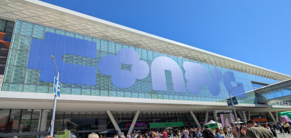 A large crowd of people is gathered outside a modern building with a glass facade. The building has a large sign with stylized letters. People are standing, talking, and taking photos. The sky is clear and blue, indicating a sunny day.
