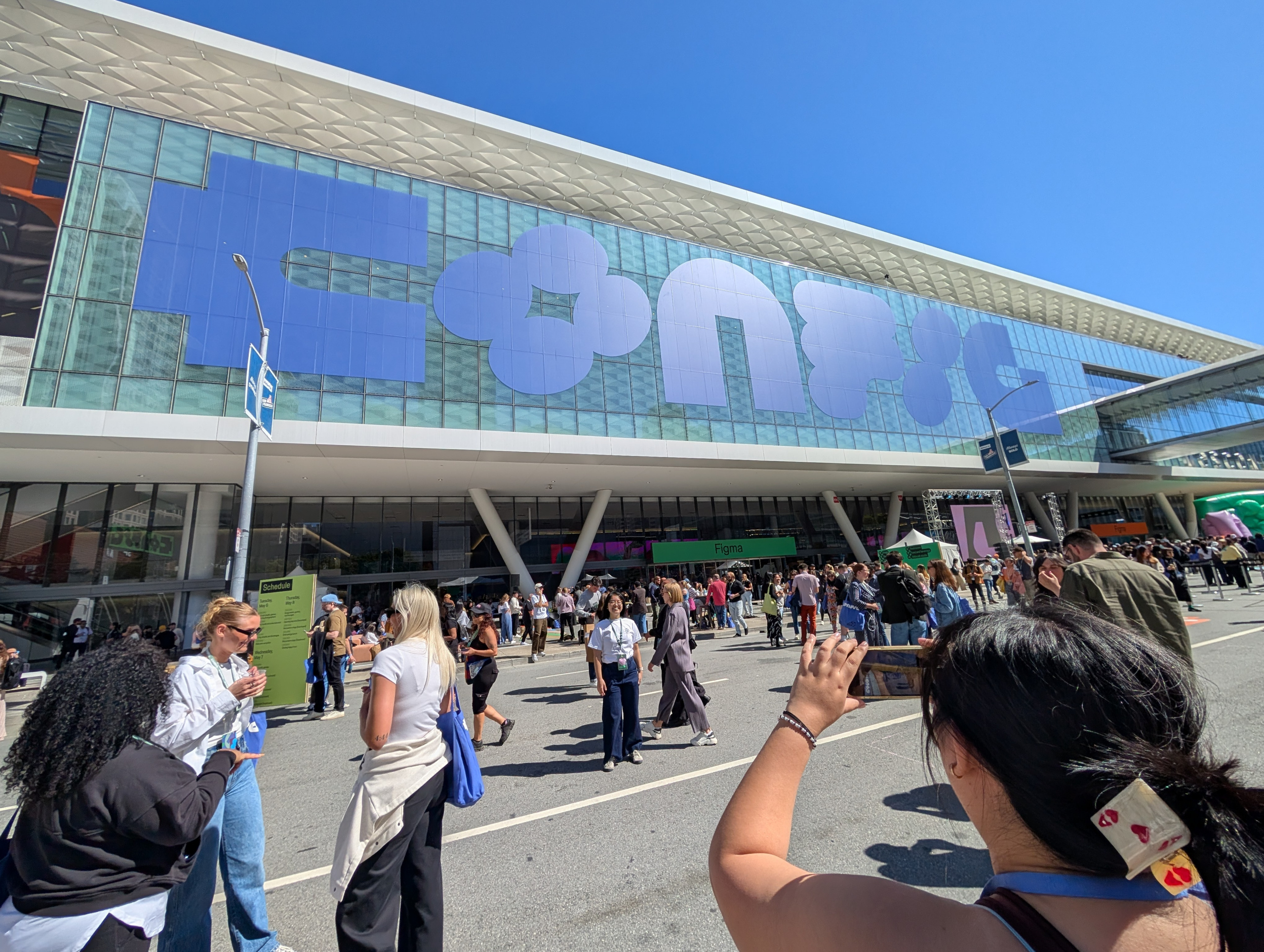 A large crowd of people is gathered outside a modern building with a glass facade. The building has a large sign with stylized letters. People are standing, talking, and taking photos. The sky is clear and blue, indicating a sunny day.